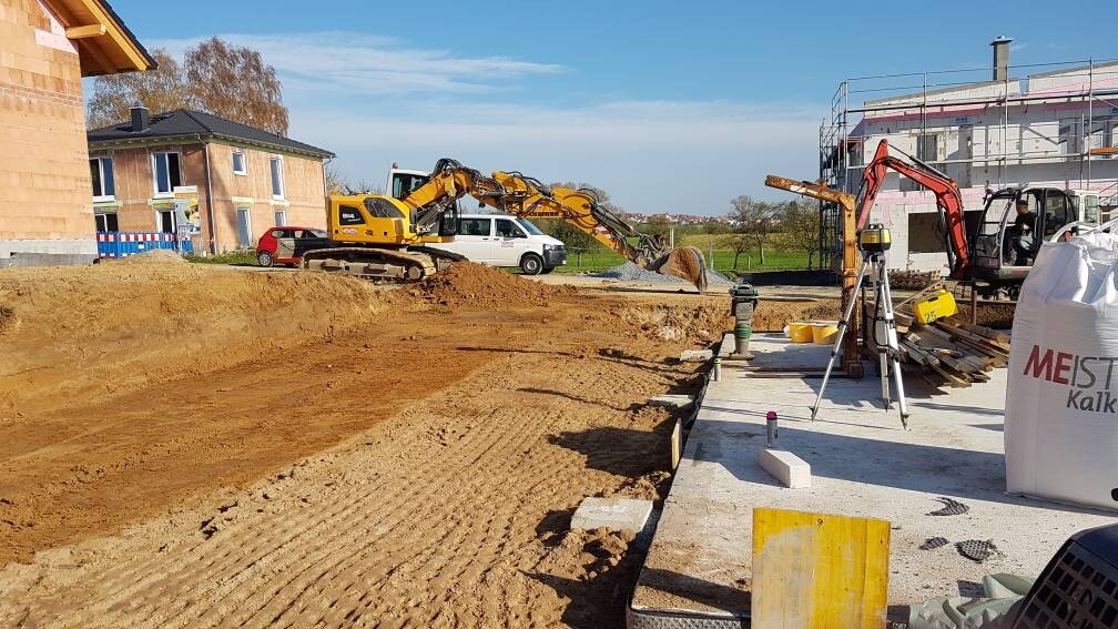 Baustelle mit Erdarbeiten: Bagger im Einsatz, Erdhaufen, Baugeräte und Rohbau im Hintergrund.