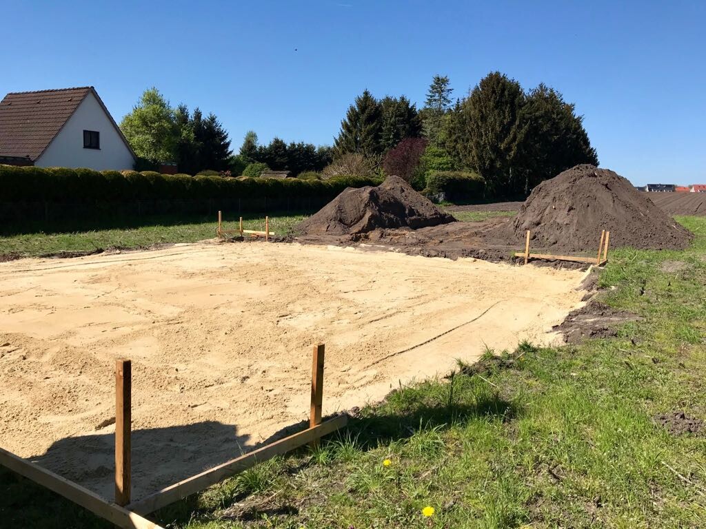 Baufläche mit Sandboden, Holzpfosten, zwei Sandhaufen; Haus und Bäume im Hintergrund.
