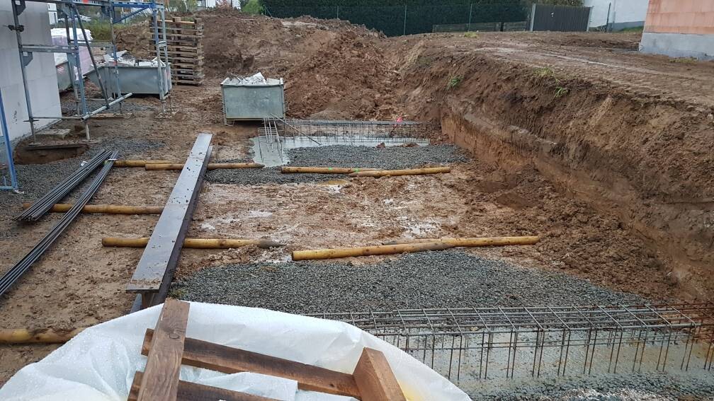 Baustelle: Fundament mit Bewehrung, Kies und Erdarbeiten; Holzbohlen liegen herum.