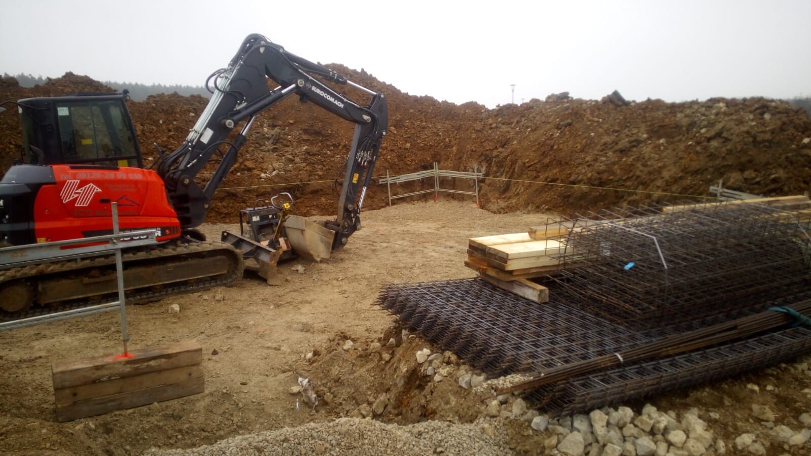 Roter Minibagger auf Baustelle neben Baugrube, Stahlbewehrung und Holzbohlen im Hintergrund