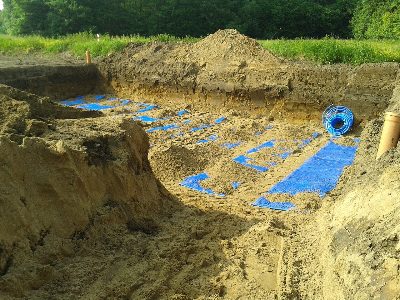 Baugrube mit blauen Schutzmatten, Rohrleitungen und Erdreich am Rand einer grünen Wiese