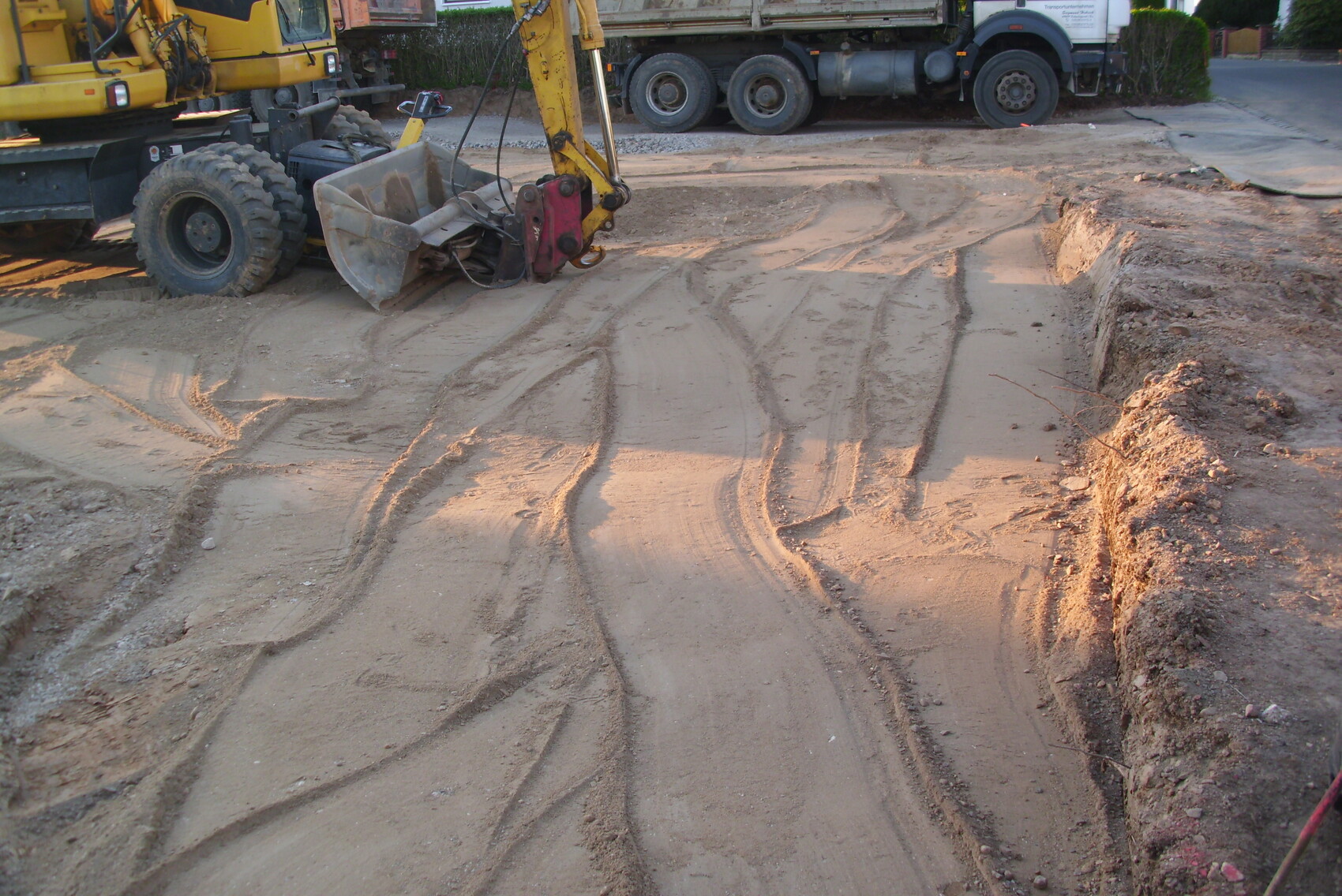 Gelber Bagger auf Baustelle; Graben mit Spuren im feuchten Sandboden.