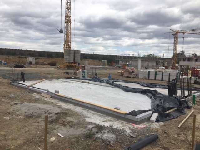 Großes Betonfundament mit schwarzen Planen auf einer Baustelle; Kräne und Baumaterial im Hintergrund.