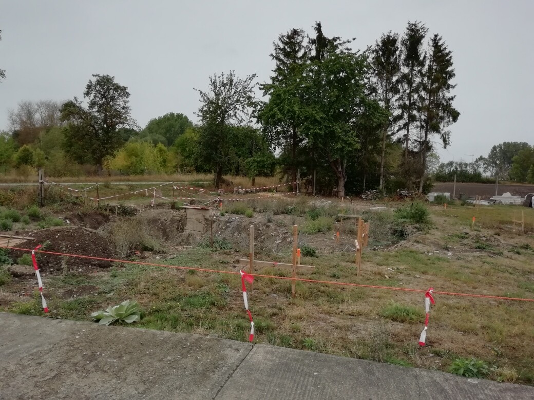 Baustelle auf unbebautem Grundstück mit Absperrband, Graben und Bäumen im Hintergrund