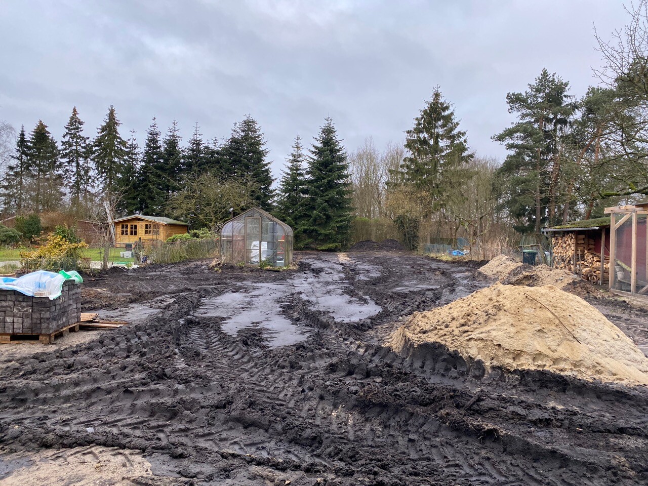 Matschige Gartenbaustelle mit Gewächshaus, Holzhaus, Sandhaufen und Baumreihe.