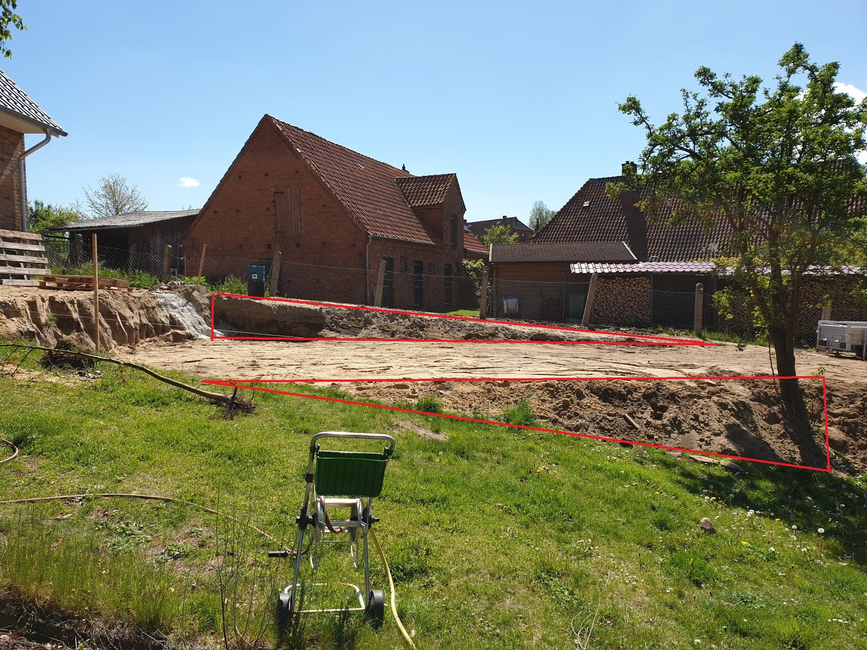 Baugrundstück mit Erdarbeiten; rote Linien markieren Gräben, Häuser im Hintergrund.