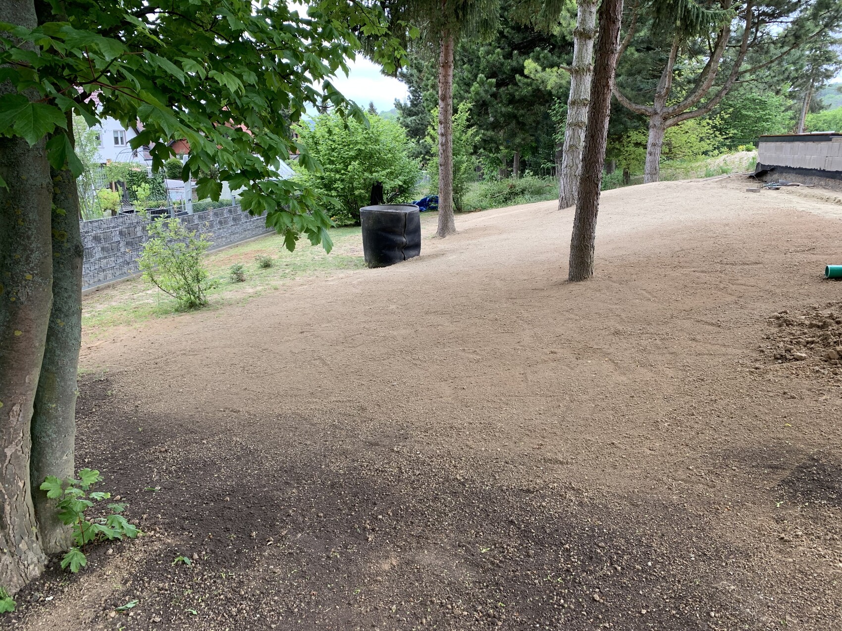 Freifläche aus Erde, Bäume, schwarzer Plastik-Tank, linke Steinmauer im Hintergrund.