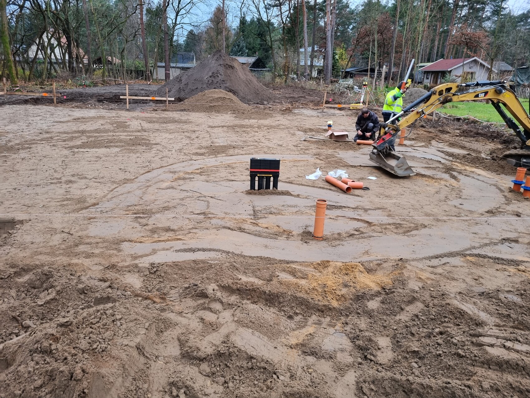 Baugrube mit Bagger, zwei Arbeiter in Warnwesten, Rohre und Sandhügel auf der Baustelle.