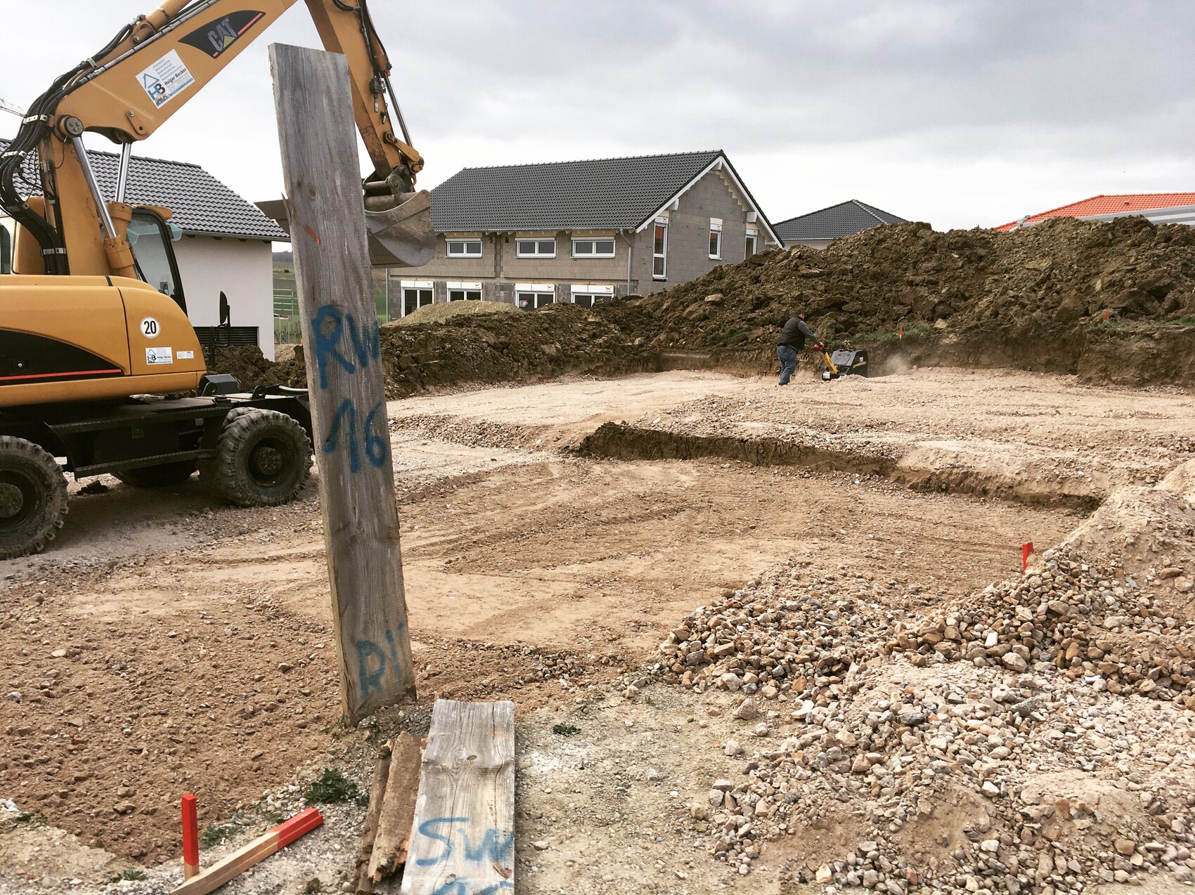 Gelber Bagger auf Baustelle, Baugrube, Arbeiter mit Verdichter, Erdehaufen, Wohnhäuser im Hintergrund.