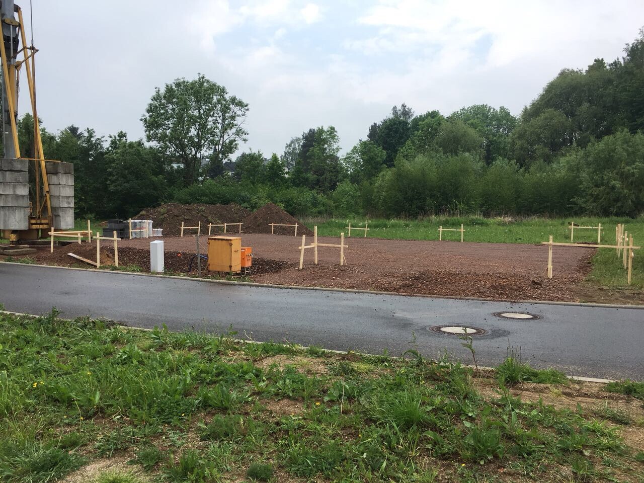 Baustelle mit freier Erdfläche, Holzpfosten-Markierungen, Baugerät links, Hintergrund mit Bäumen.
