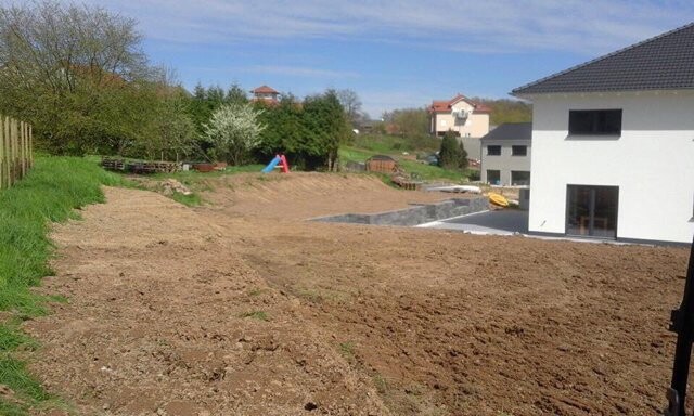 Baustelle im Garten eines modernen Hauses; Erdarbeiten, Baufläche und Spielgeräte im Hintergrund.