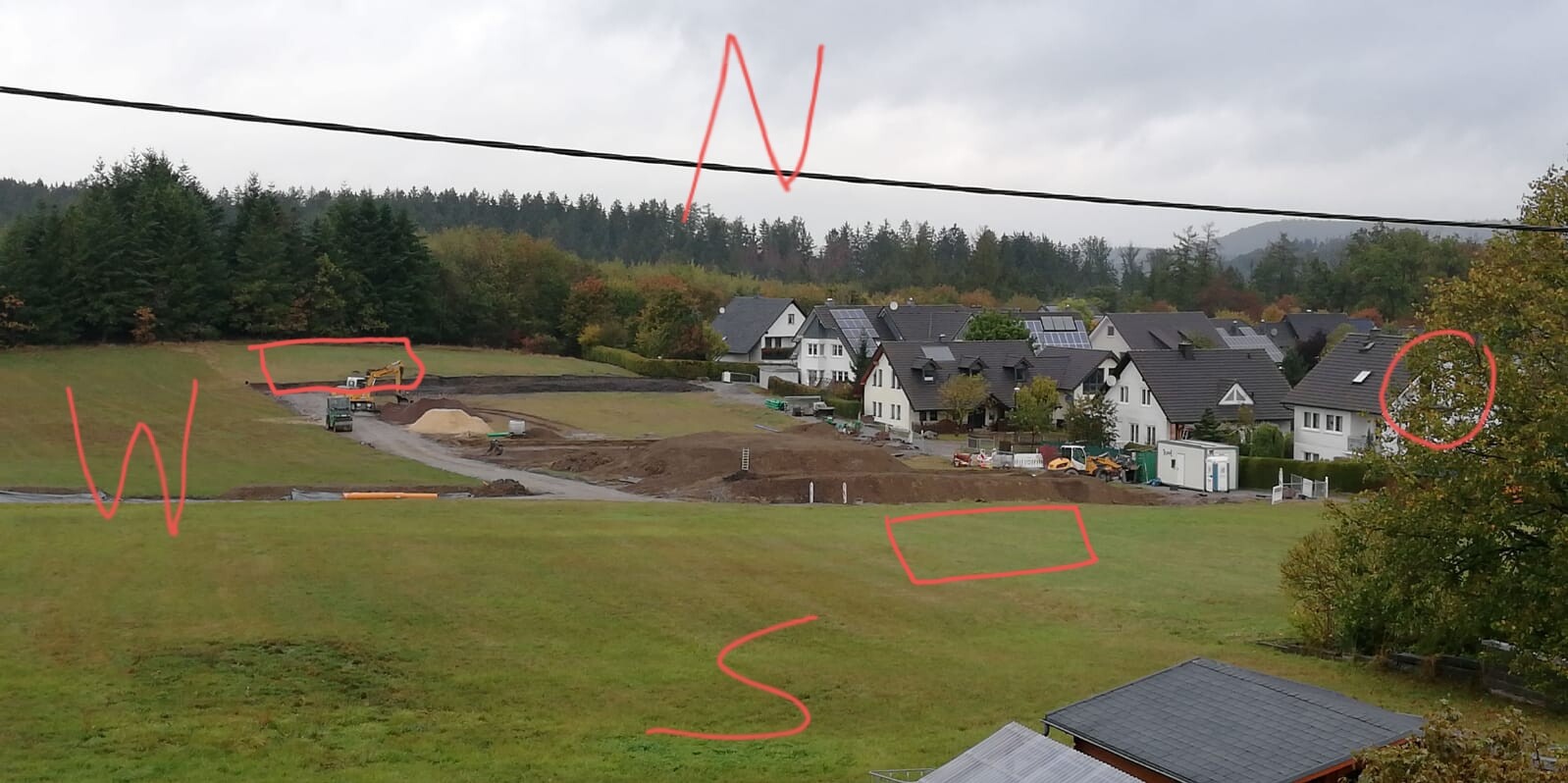 Baustelle vor Wohnsiedlung mit Bagger, Erdhaufen und Häusern im Hintergrund.