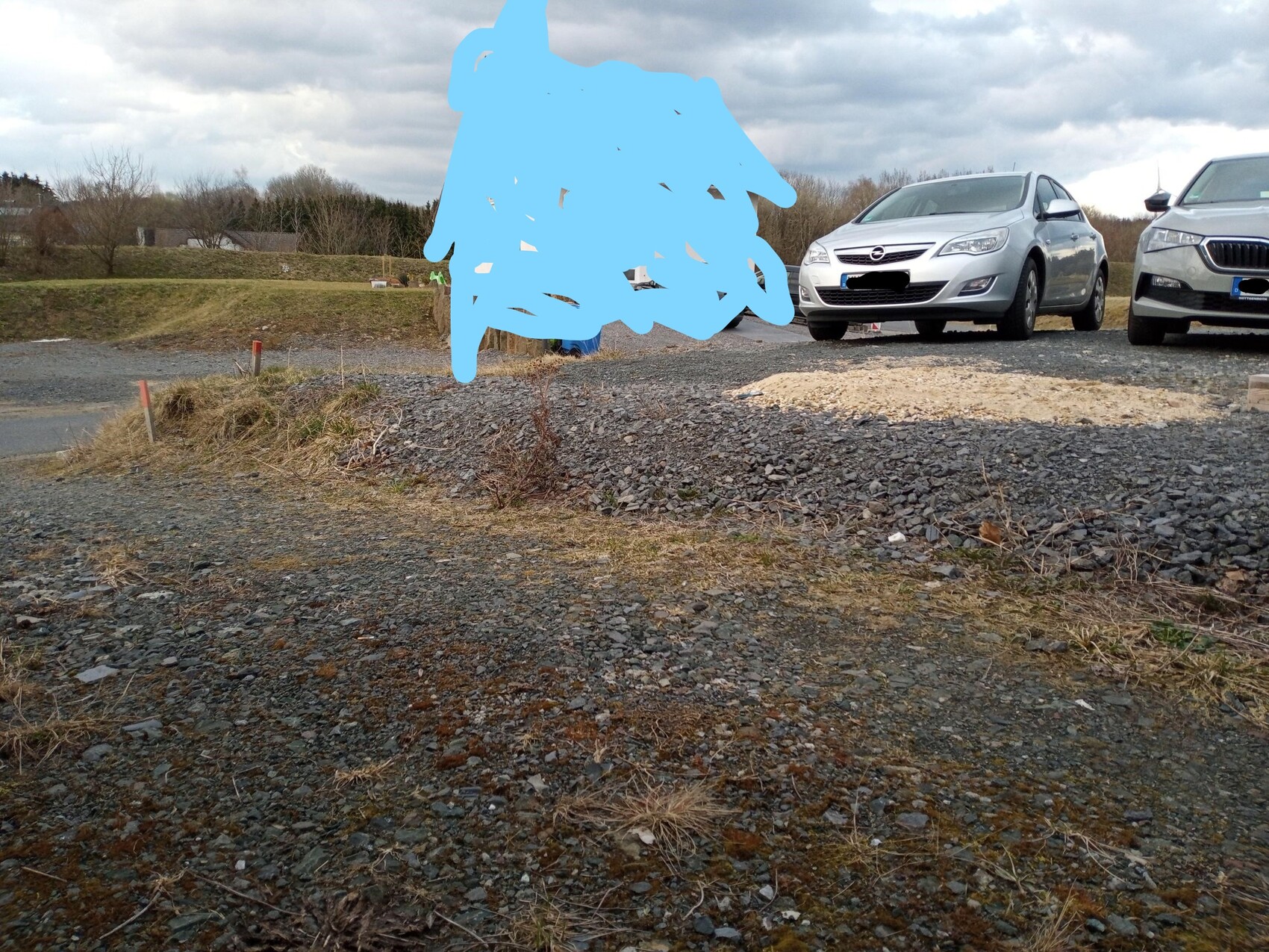 Parkplatz mit zwei Autos rechts, Kiesboden; blaue Markierung verdeckt einen Teil der Szene.