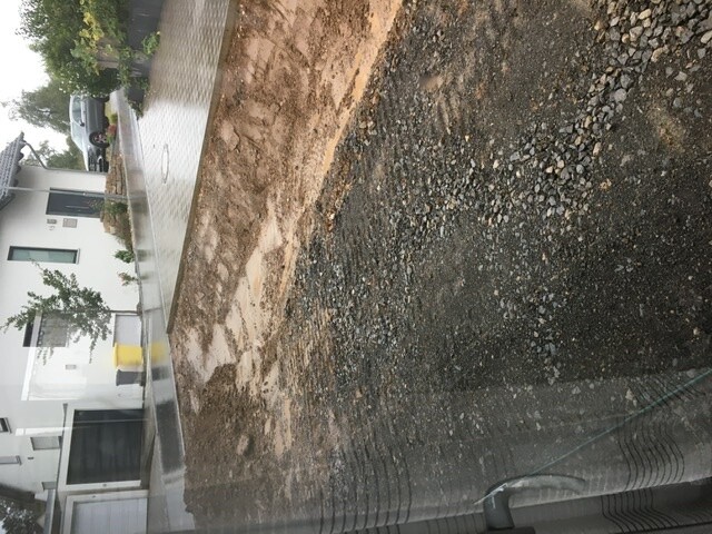 Baustelle neben Wohnhaus: unbefestigte Erde und Kies, linke Straßenseite nass.