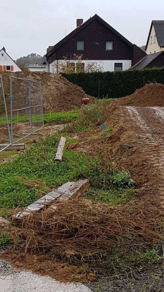 Baustelle mit Erdhaufen, Holzresten und Zaun, Wohnhaus im Hintergrund.