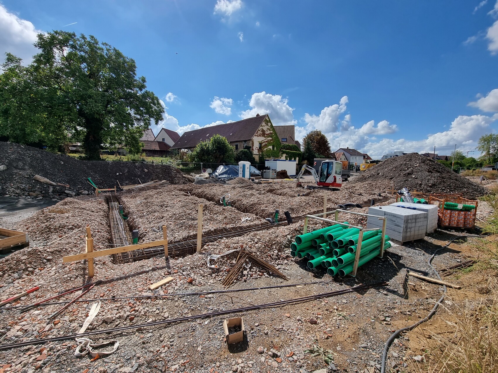 Baustelle in Vorstadt: Erdhaufen, grüne Rohre, Bagger, Bauholz und Häuser im Hintergrund.