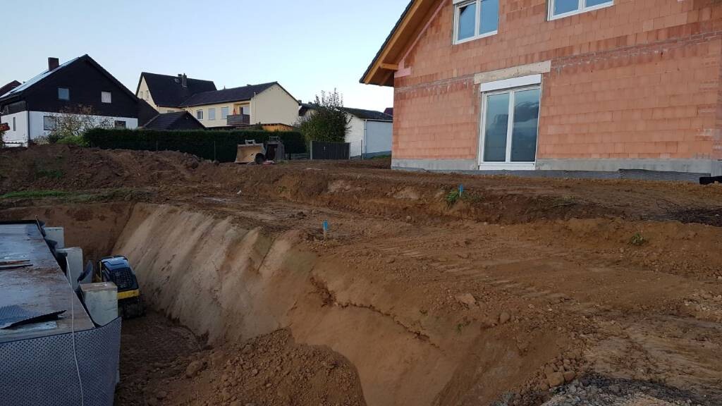 Baustelle mit tiefem Erdgraben neben unfertigem Haus; Nachbarschaft im Hintergrund.