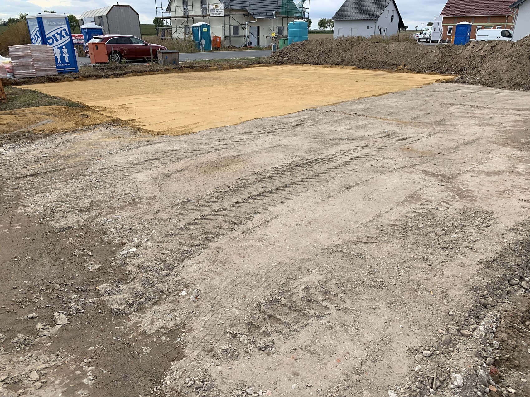 Baustelle mit Erdarbeiten: Sandfläche vorbereitet, Bauequipment sichtbar, Häuser im Hintergrund.