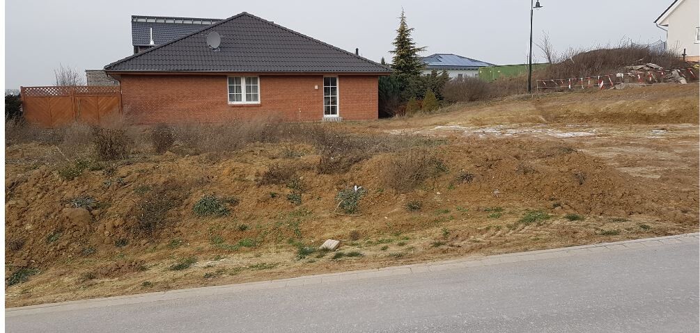 Rotes Einfamilienhaus mit dunklem Dach vor trockenem Baugelände und Straße im Vordergrund.