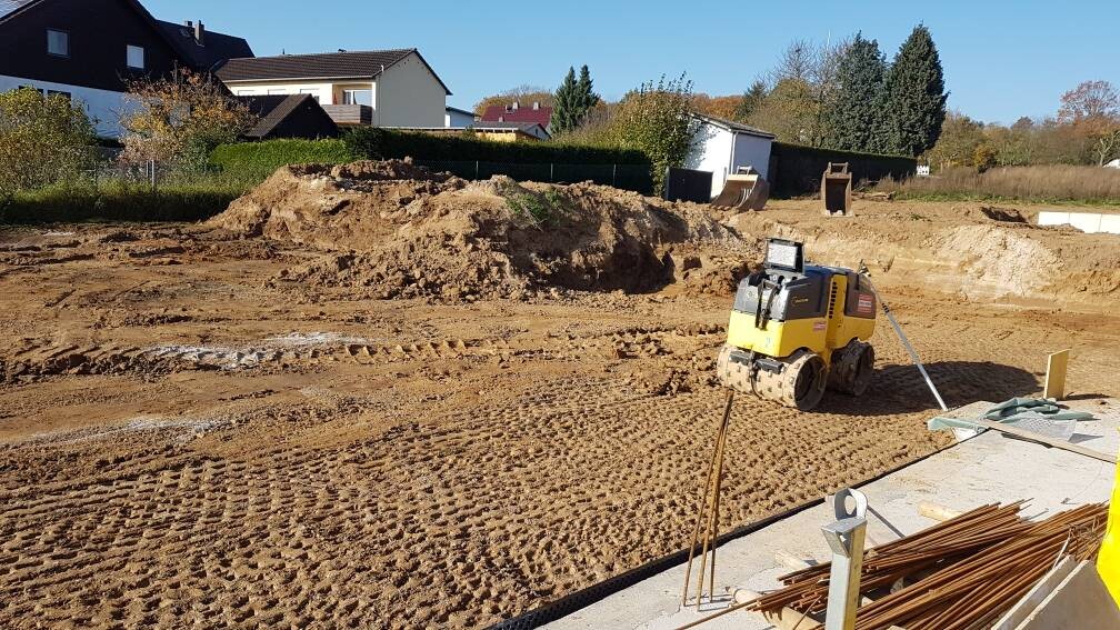 Auf einer Baustelle: Gelber Kompaktlader, Erdaushub, Stahlstangen, Bauholz, Häuser im Hintergrund.