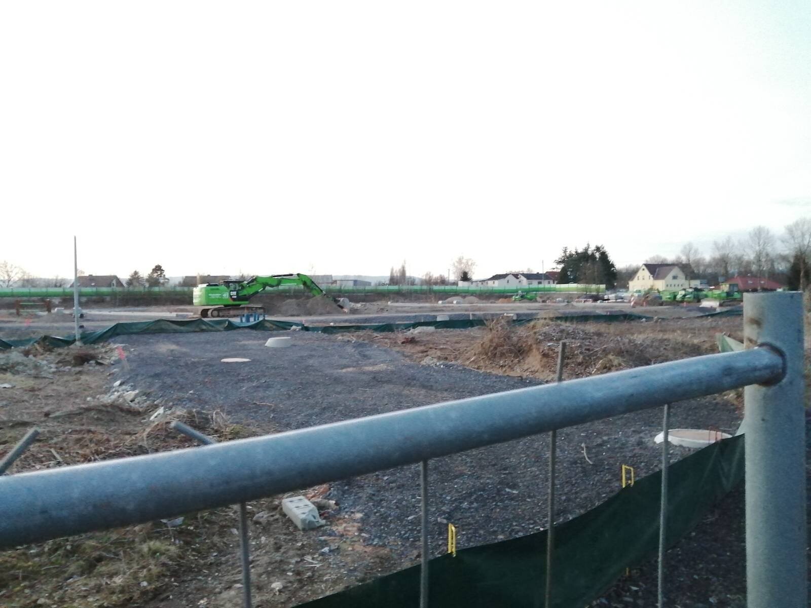 Baustelle mit grünem Bagger, Erdaushub, Bauzäune; Häuserreihe im Hintergrund