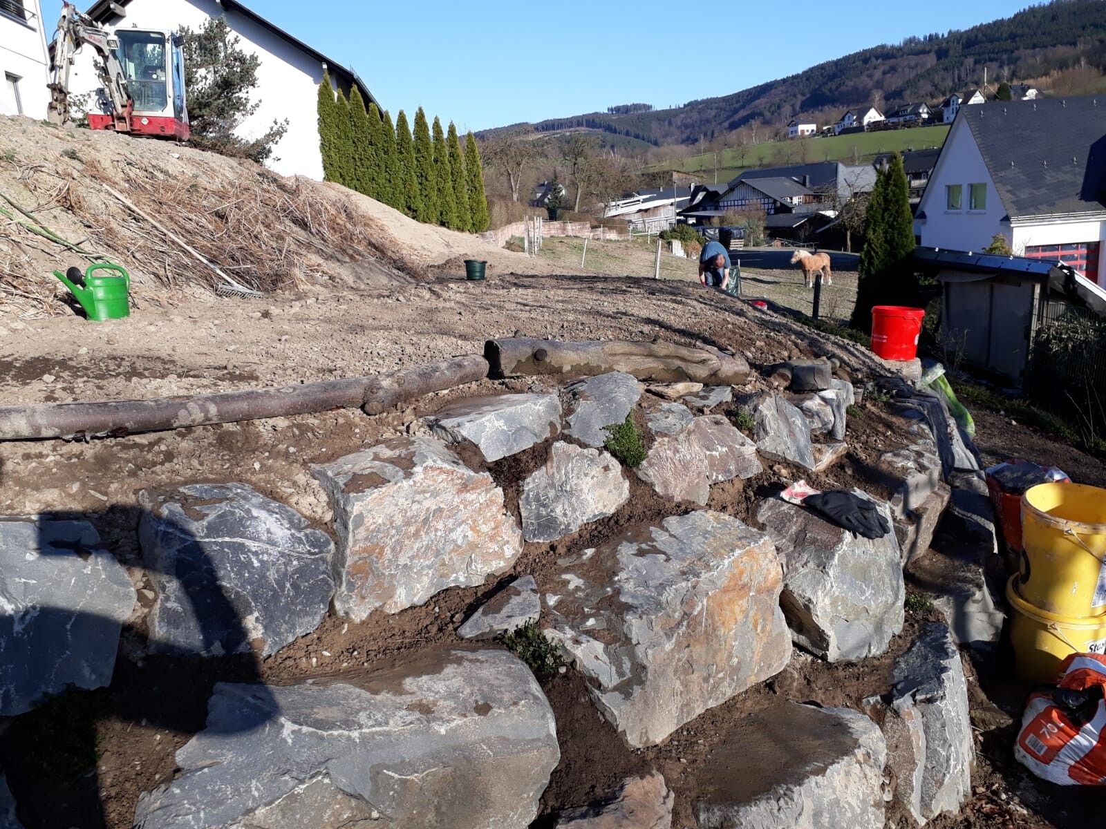 Trockenmauer aus großen Steinen am Hang; Bagger links, Eimer vor der Mauer, Häuser im Hintergrund.