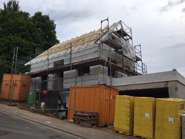 Rohbau eines Hauses mit Gerüst, sichtbarer Dachkonstruktion, orange Container und gelbem Dämmstoff.