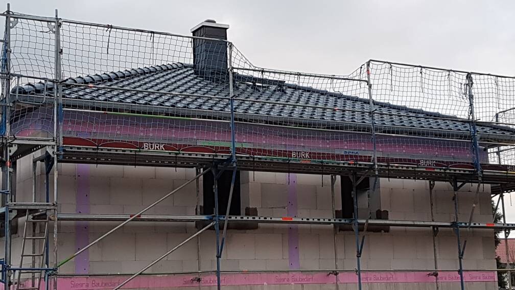 Hausbaustelle mit Gerüst, Netzen, dunklem Ziegeldach und pinken Fassadenplatten.