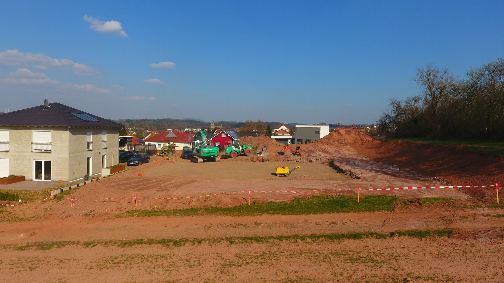 Baustelle vor Wohnsiedlung: neue Baugrube, Bagger, Erdehaufen, Absperrband, links Haus.