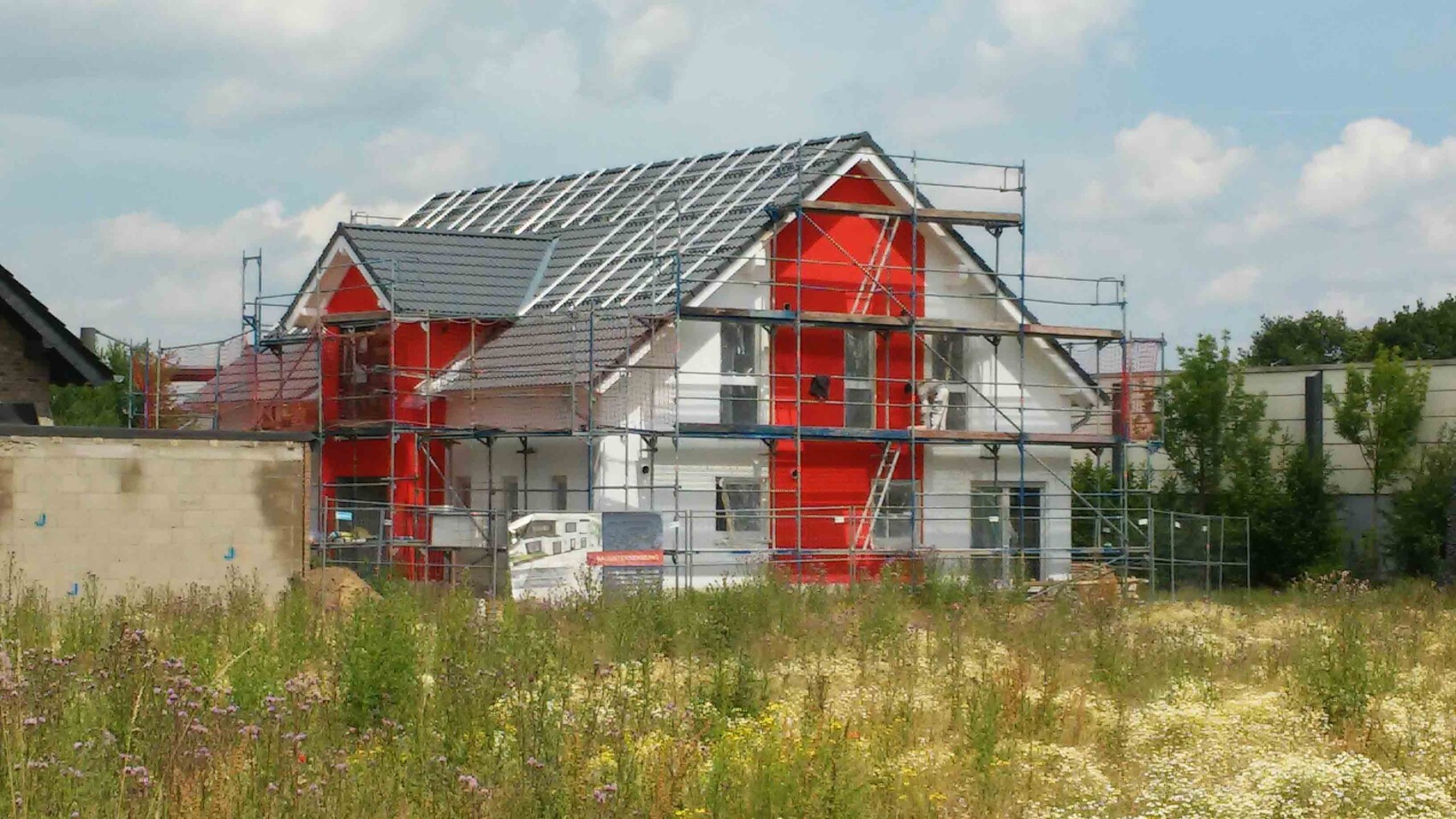 Haus im Rohbau mit Gerüst, rote Wandflächen, sichtbarer Dachstuhl, Wiese davor