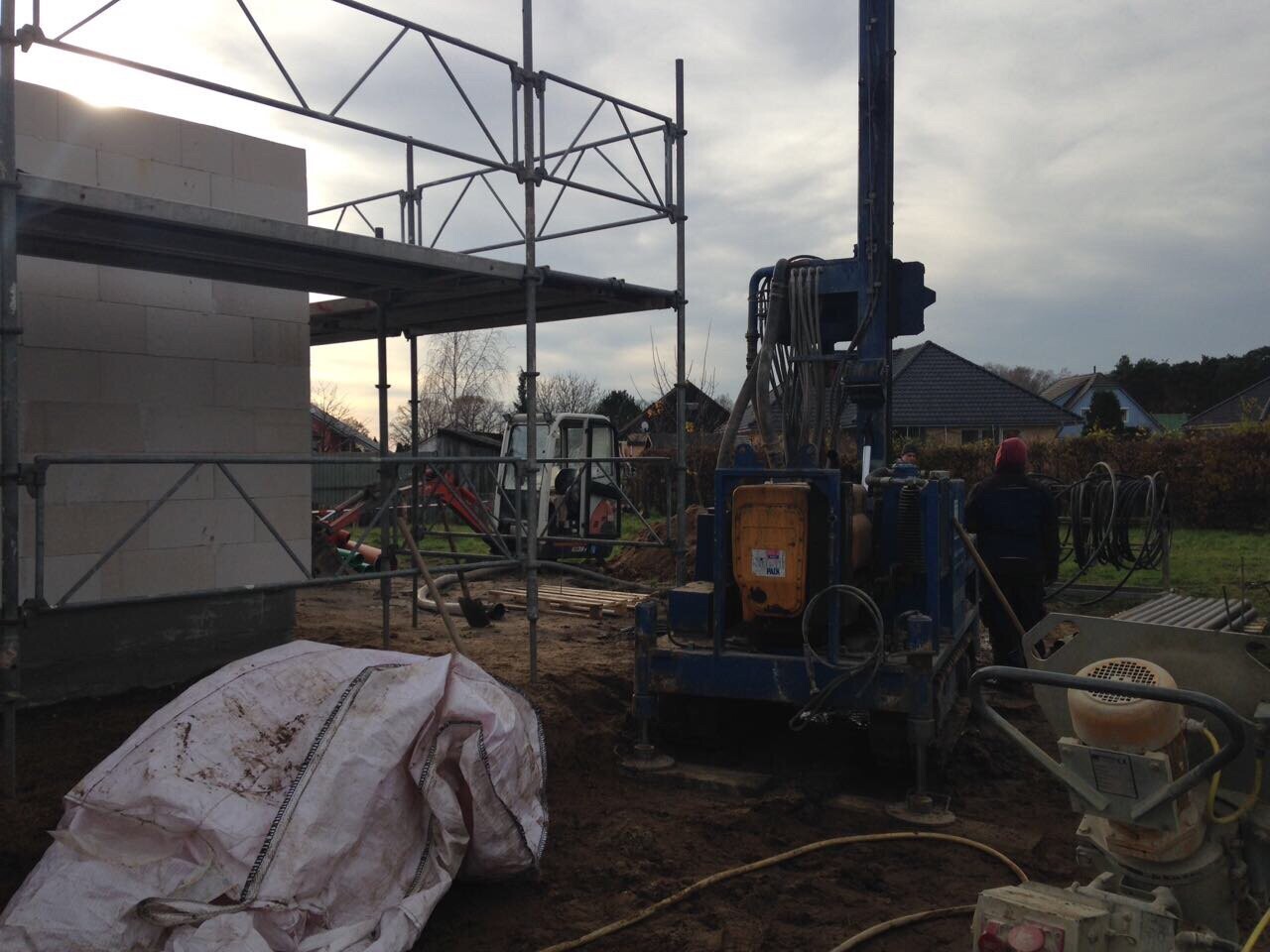Große Bohranlage und Gerüst auf Baustelle neben Wohnhaus, Schlamm, Kabelrollen und Baumaterialien.