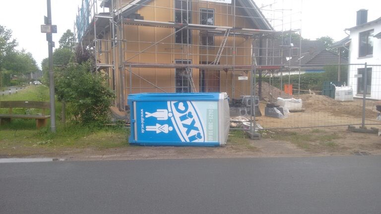 Baustelle mit Gerüst am Haus; blaue tragbare Toilette liegt auf dem Gehweg.