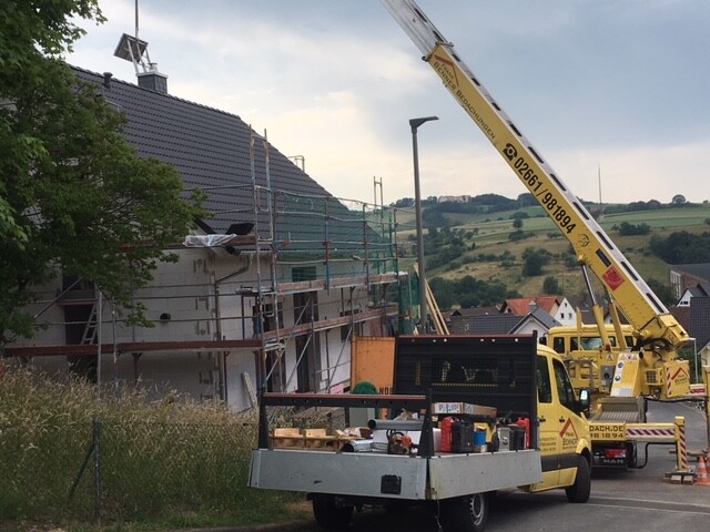 Baustelle mit Gerüst um ein Wohnhaus, Kranwagen und beladener Anhänger vor ländlicher Umgebung.