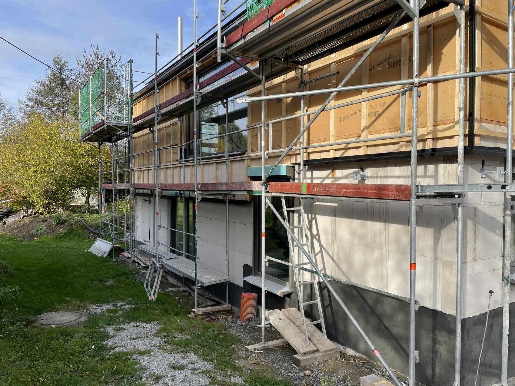Außenfassade eines Gebäudes im Bau mit Holzverkleidung und umlaufendem Metallgerüst, Gartenbereich.