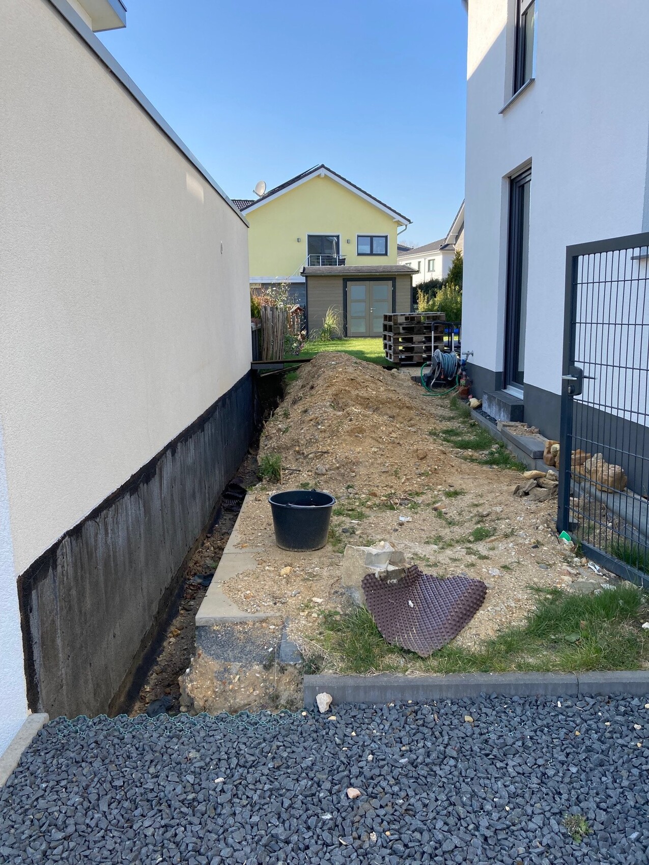 Gartenbaubaustelle zwischen weißen Häusern: Erdhügel, Eimer, Paletten, Graben und Kies.