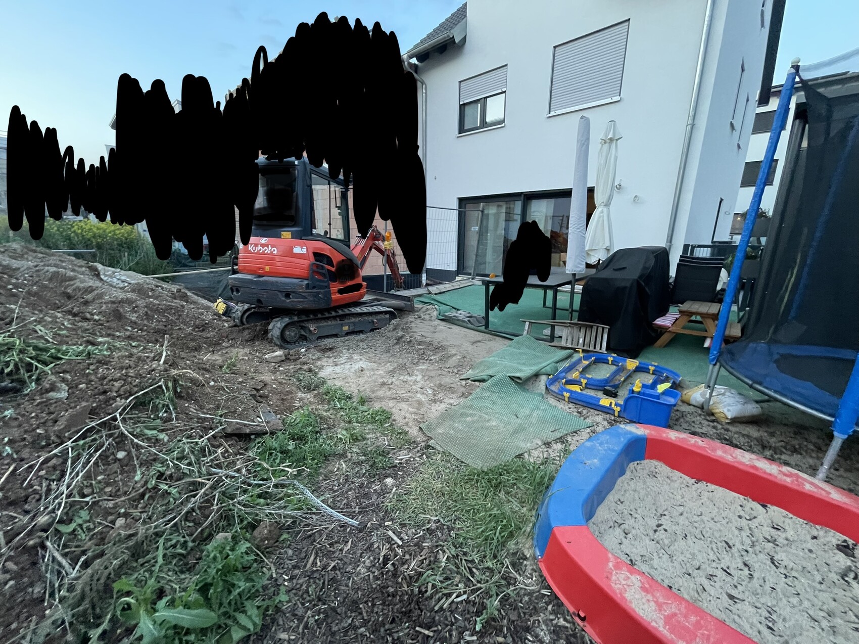 Kleiner roter Minibagger arbeitet an Baugrund vor modernem weißen Haus mit Terrasse und Gartenzubehör