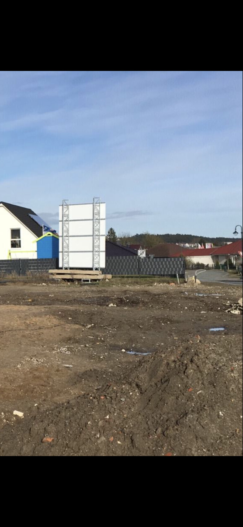 Baustelle mit leerer Werbetafel auf Metallrahmen vor Wohnhäusern; unbefestigter Boden.