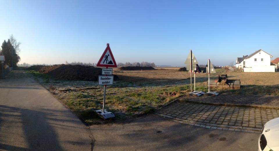 Straßenbaustelle in einem Wohngebiet mit Erdmassen, Schild und Häusern im Hintergrund.