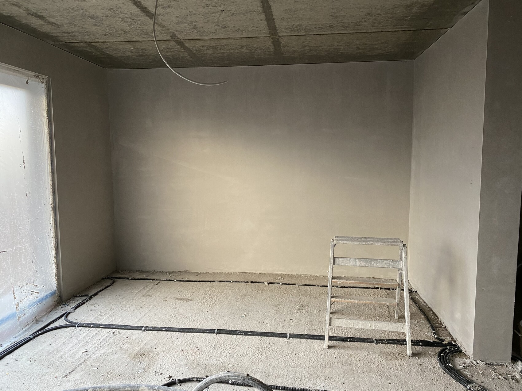 Unverputzter Bau-Raum: Leiter, Kabel am Boden, rauer Beton, offene Deckenleitungen.