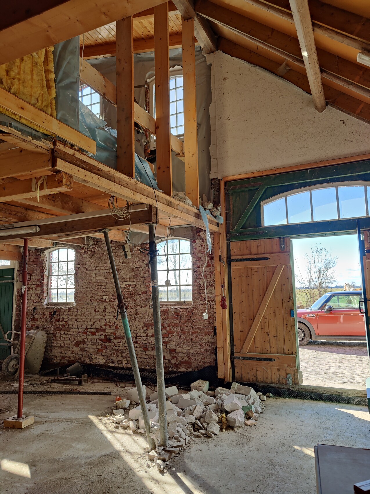 Baustelleninterieur: freigelegtes Ziegelmauerwerk, Holzträger, Trümmer, offenes Tor mit Auto draußen.
