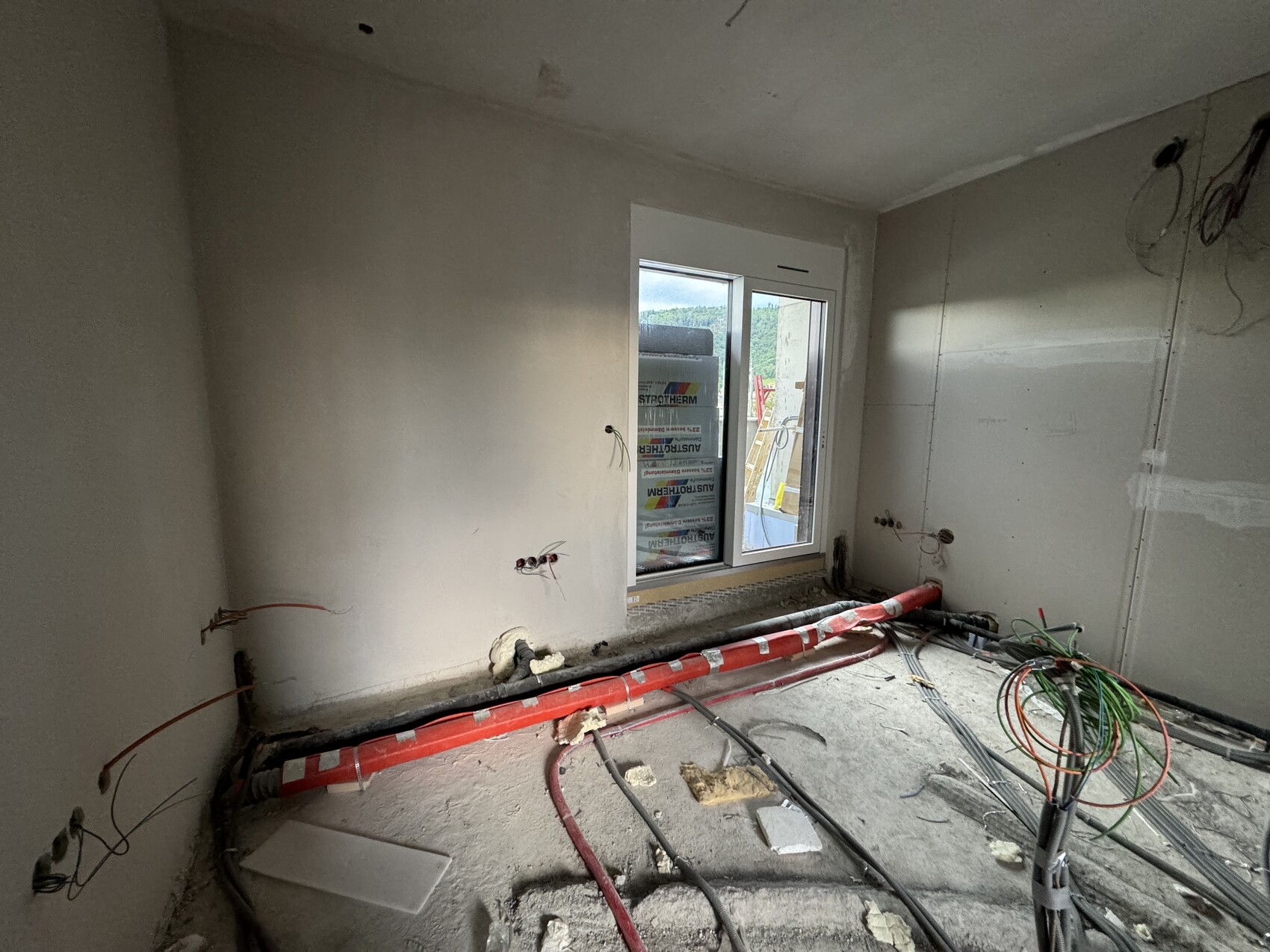Renovierungsraum mit freiliegenden Rohren, Kabeln und Fenster zum Außenbereich.