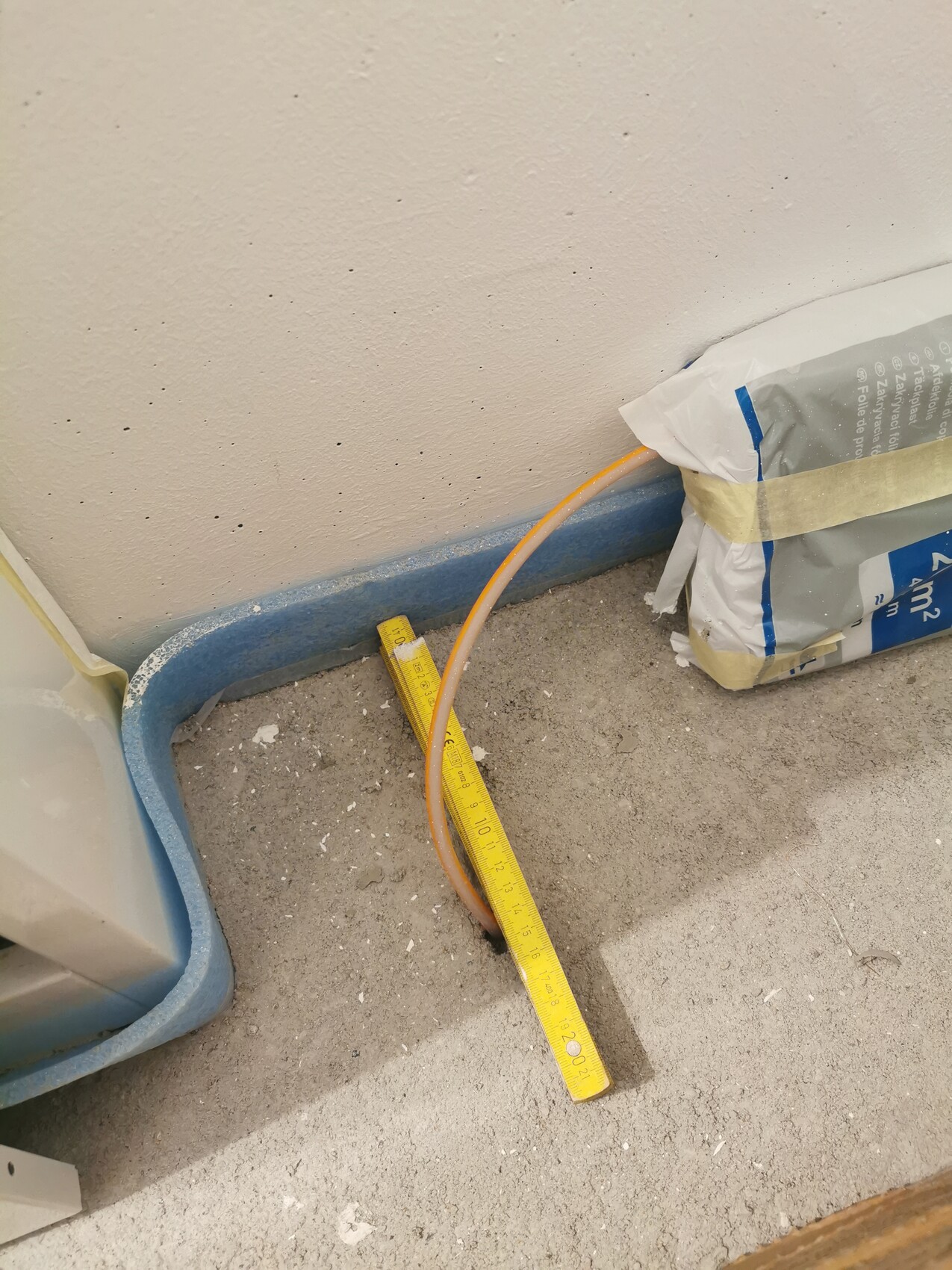 Baustelle: offenes Bodenloch mit Messband darüber, orange Schlauch, rechts Baustoffbeutel.