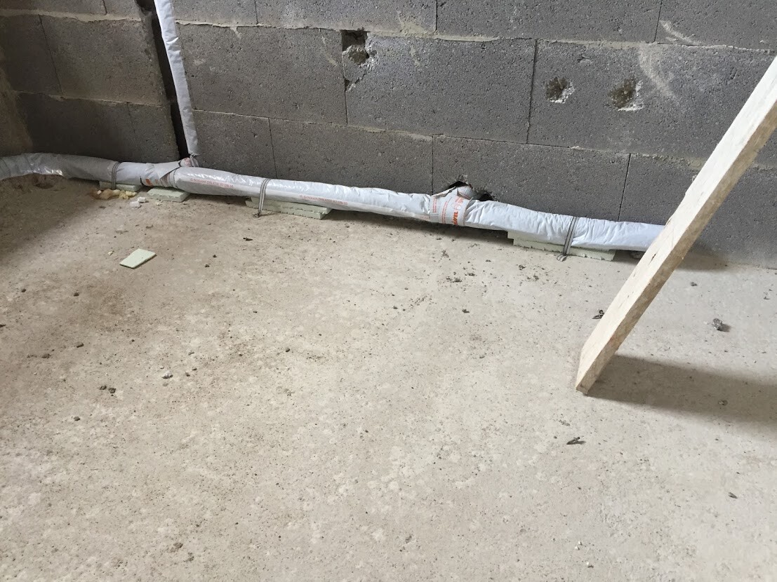 Kellerbaustelle: Betonwand mit Löchern, isoliertes Rohr verläuft am Boden, rechts eine Holzlatte.