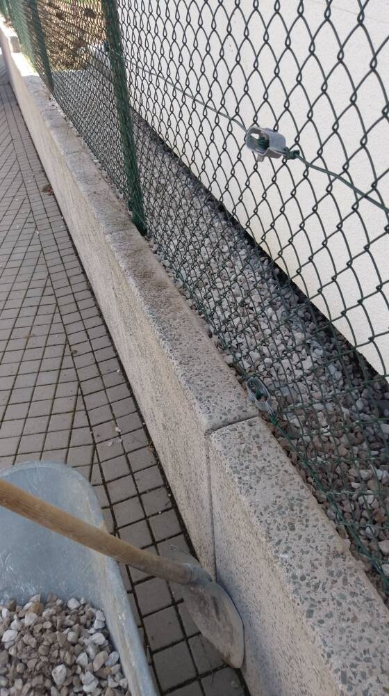 Drahtgitterzaun neben Betonbordstein, Kies dazwischen; vorn eine Schubkarre mit Schaufel.