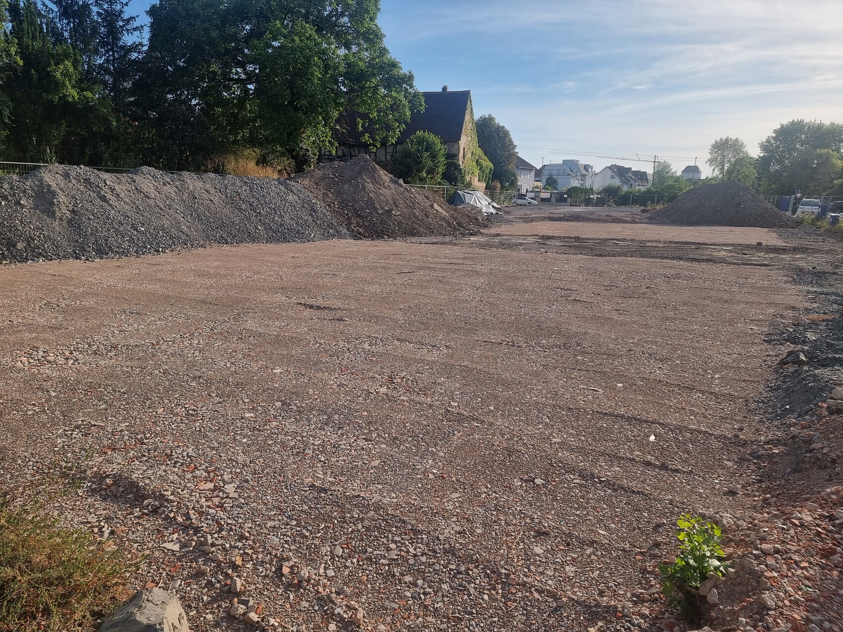 Großes unbefestigtes Baugrundstück mit Kieshaufen links und rechts; Häuser im Hintergrund.