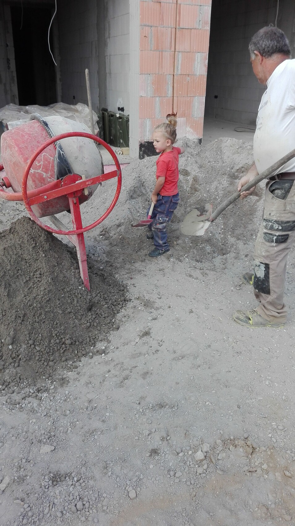 Junge in rotem T?Shirt neben rotem Betonmischer; Bauarbeiter schaufelt Kies auf Baustelle.