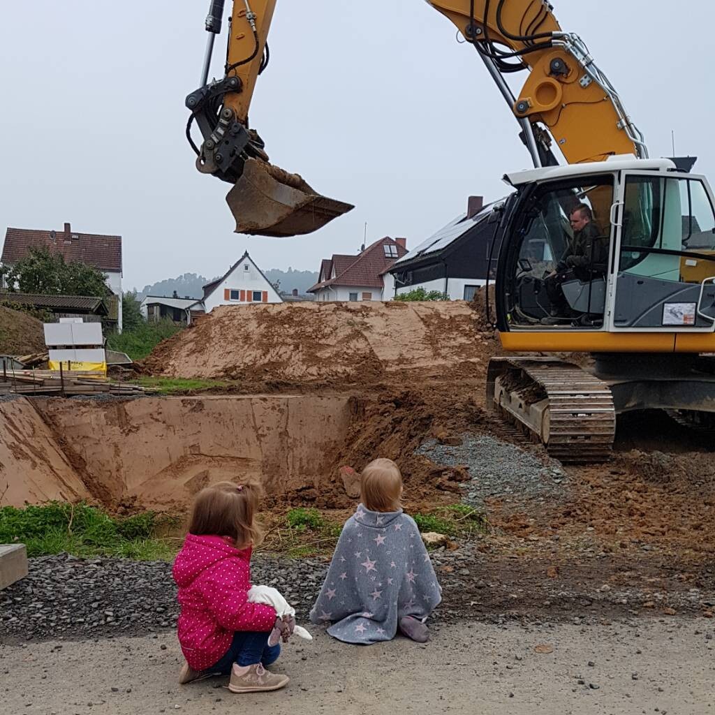 Zwei Kinder sitzen am Baustellenrand und beobachten einen gelben Bagger beim Graben.