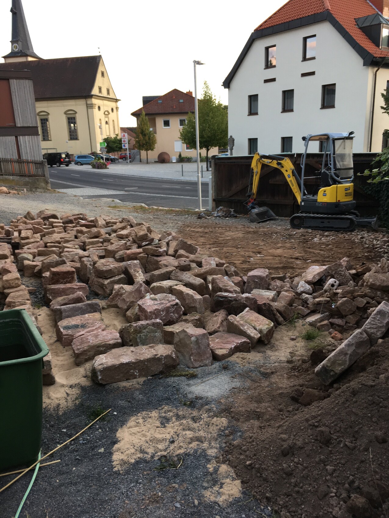 Baustelle in einer Stadt mit Steinblöcken, Minibagger, Straßen und Wohnhäusern im Hintergrund.
