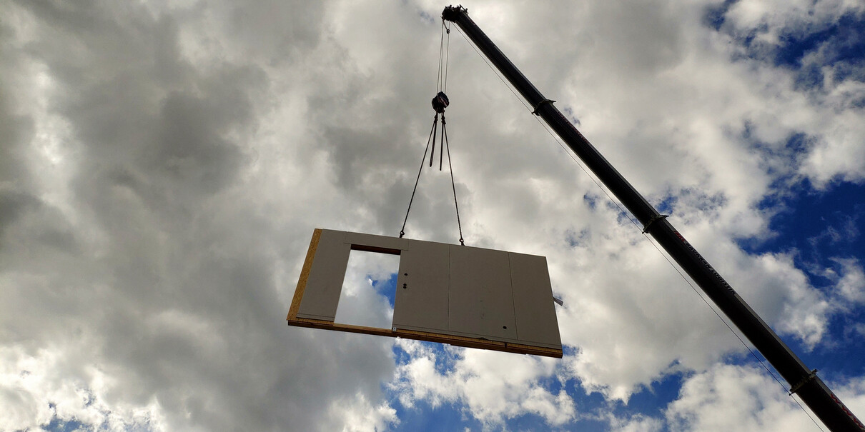 Kran hebt eine große Wandplatte mit Fensteröffnung vor wolkigem Himmel.