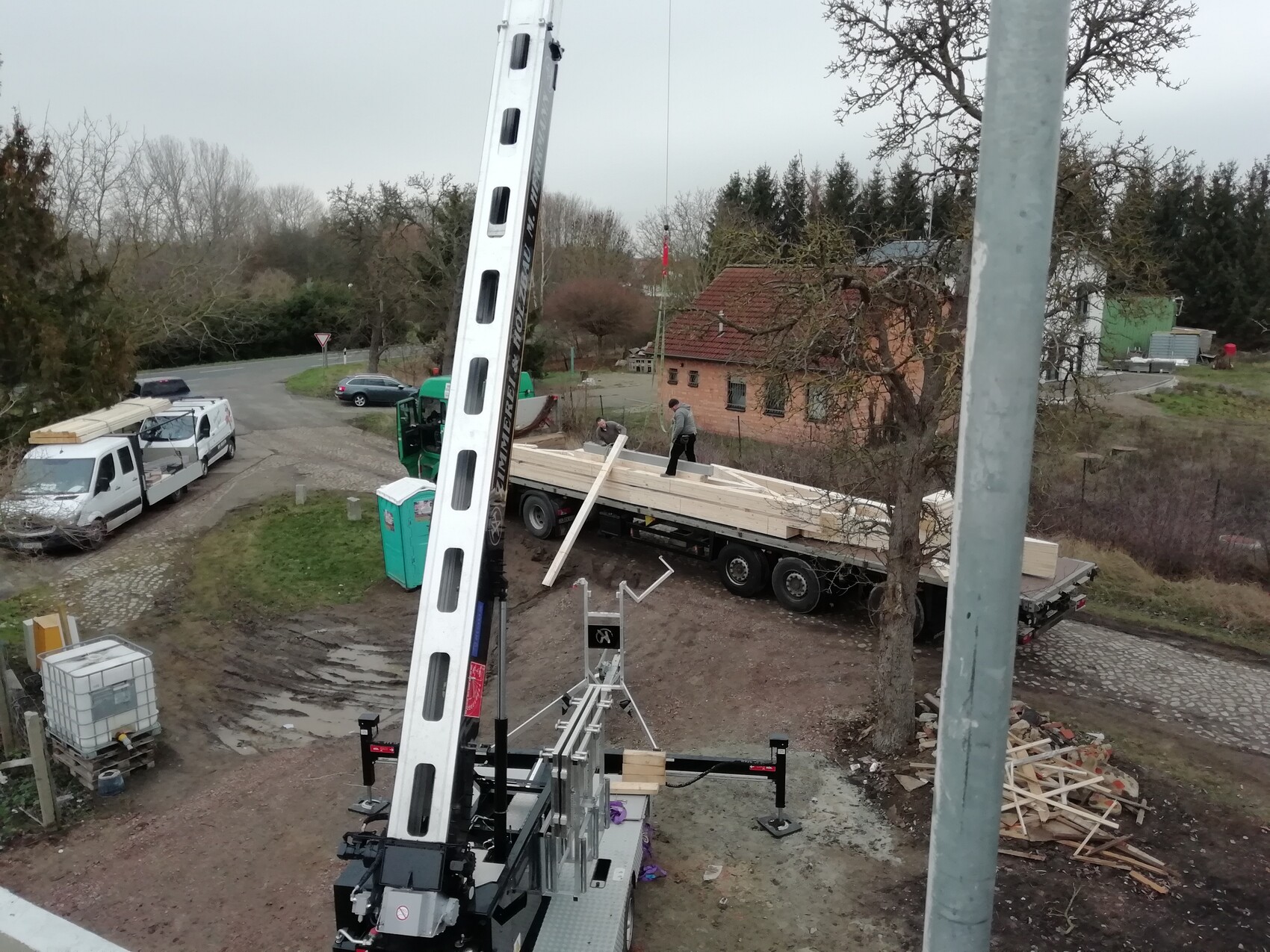Baustelle mit Kran an Lastwagen, lange Holzbalken auf dem Trailer; zwei Arbeiter arbeiten.